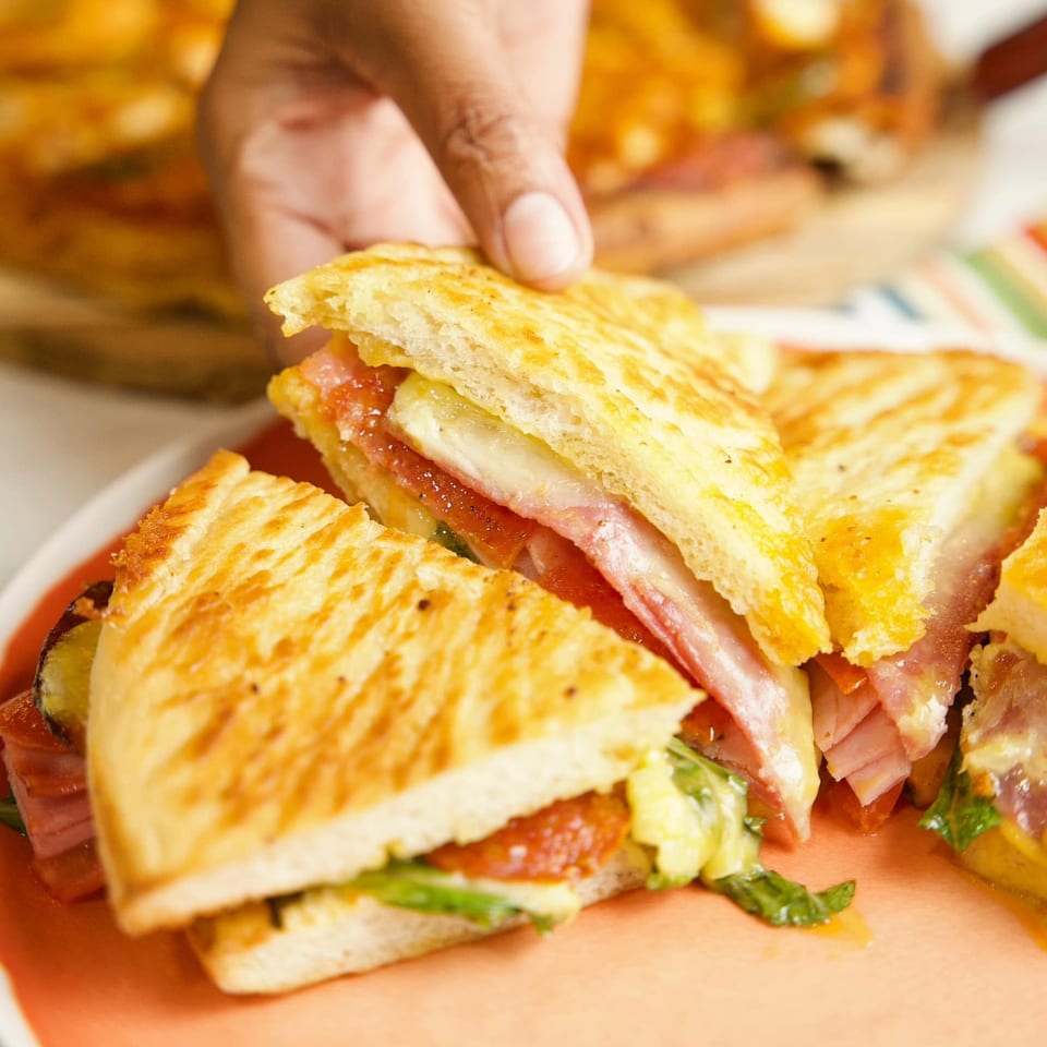 Hand picking up a golden-brown grilled sandwich filled with ham, cheese, and vegetables.
