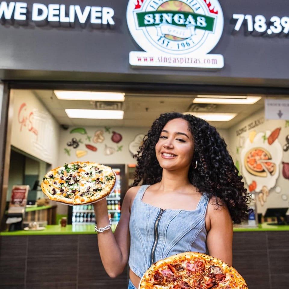 Smiling woman holding two pizzas in front of a Singas Famous Pizza restaurant.