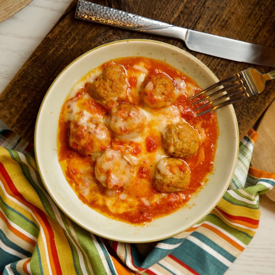Meatballs in tomato sauce with melted cheese, a fork, and a colorful striped napkin.