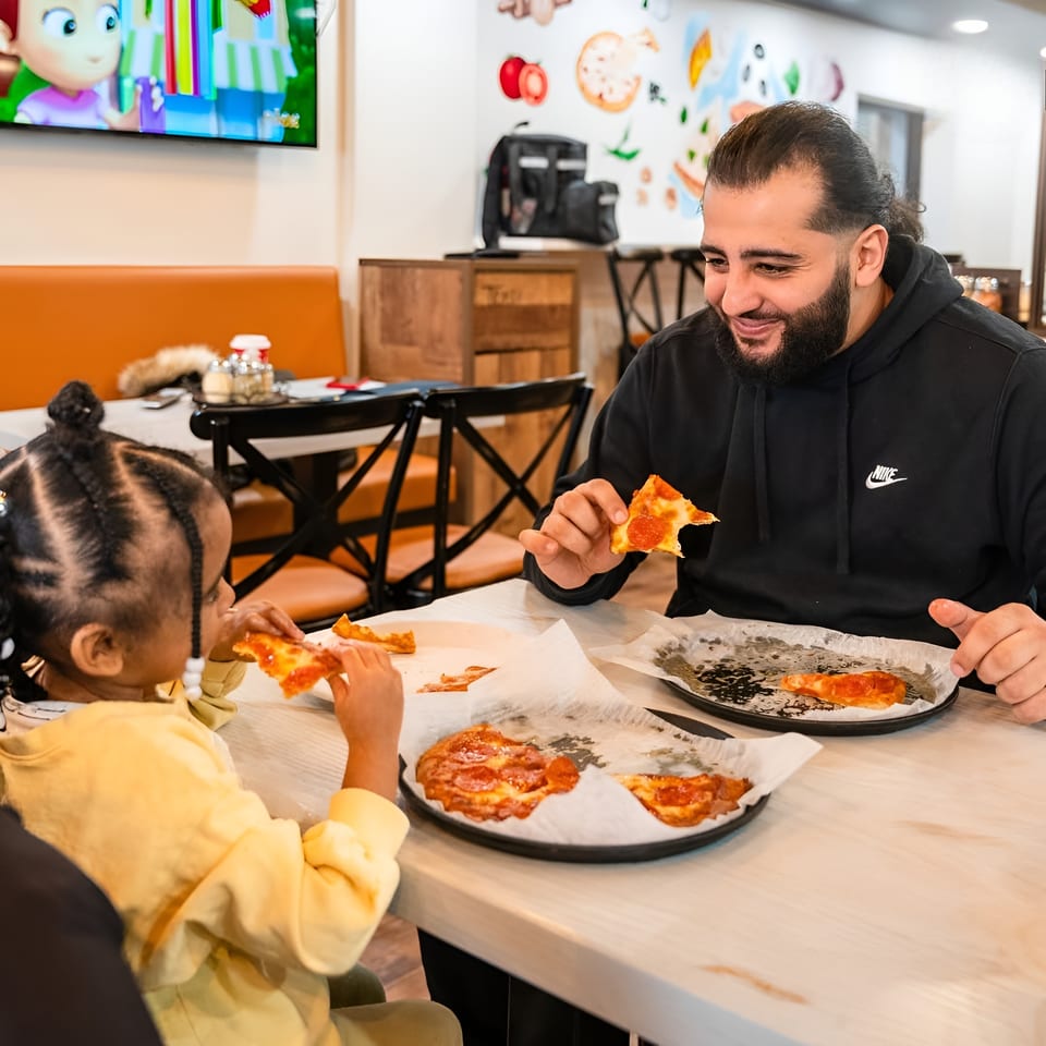 A man and a child eating pizza at a table in a brightly lit restaurant.