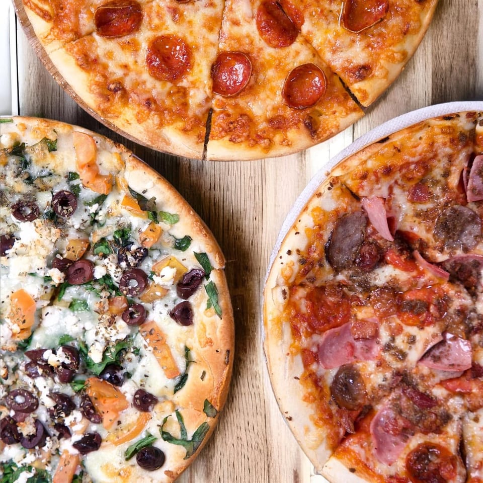 Three different pizzas, including pepperoni, veggie, and meat lover's, on a wooden table.
