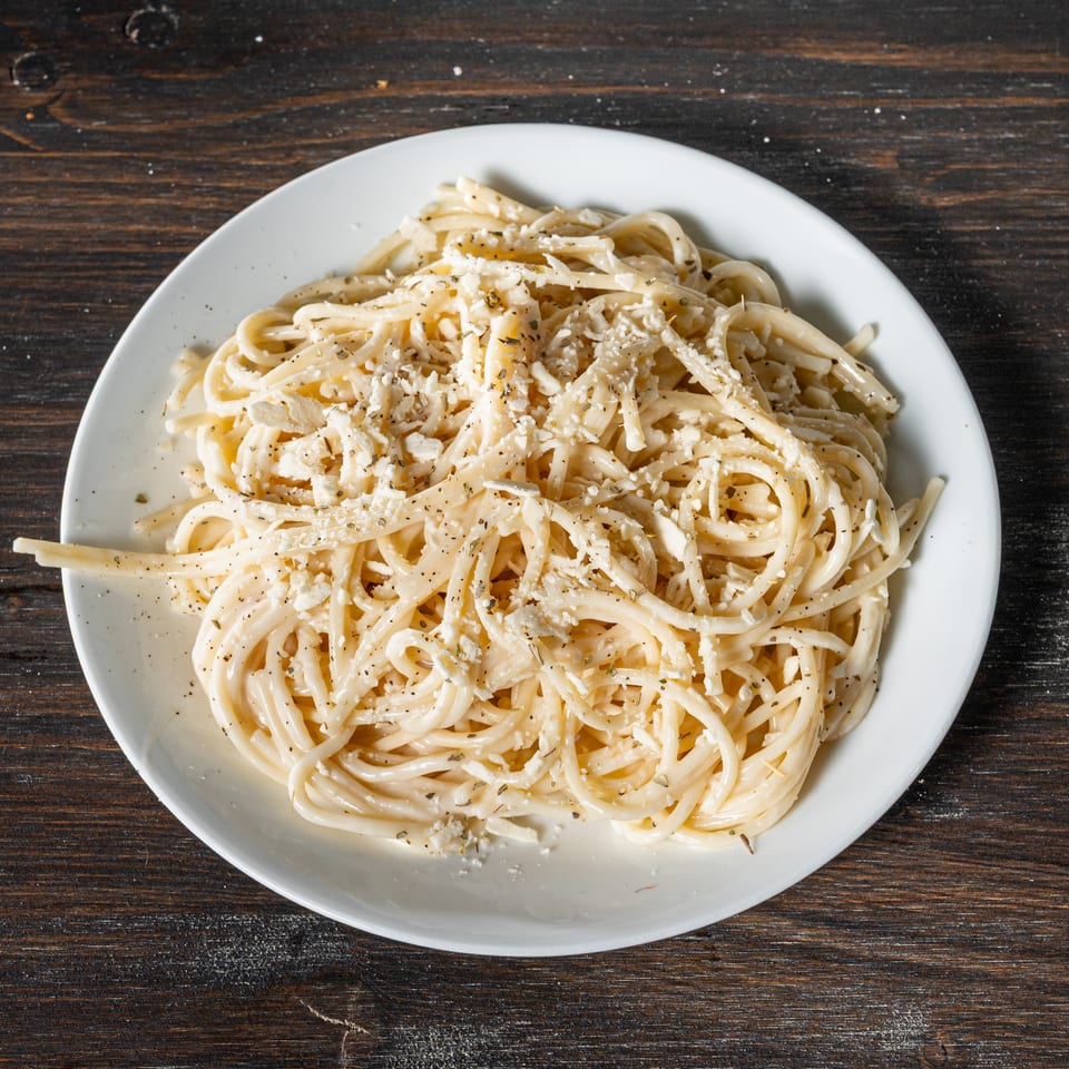 A white plate of spaghetti with white sauce, cheese, and black pepper on a dark wooden table.