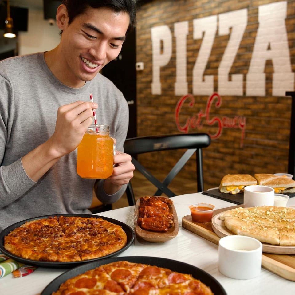 Smiling man enjoying a drink at a table filled with pizzas, wings, and sandwiches.