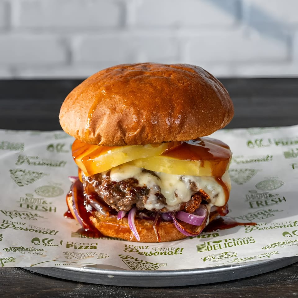 Gourmet burger with melted cheese, pineapple, red onion, and BBQ sauce on a brioche bun.