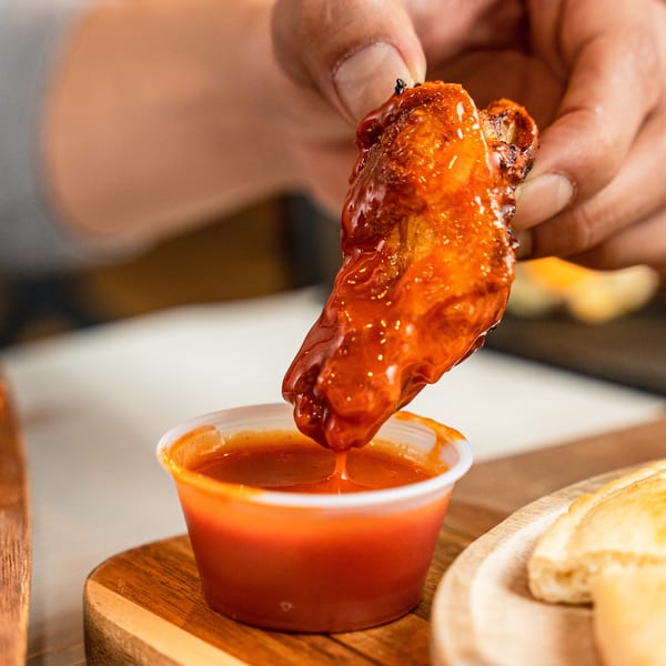 A hand dips a glistening, saucy chicken wing into a small cup of red sauce.