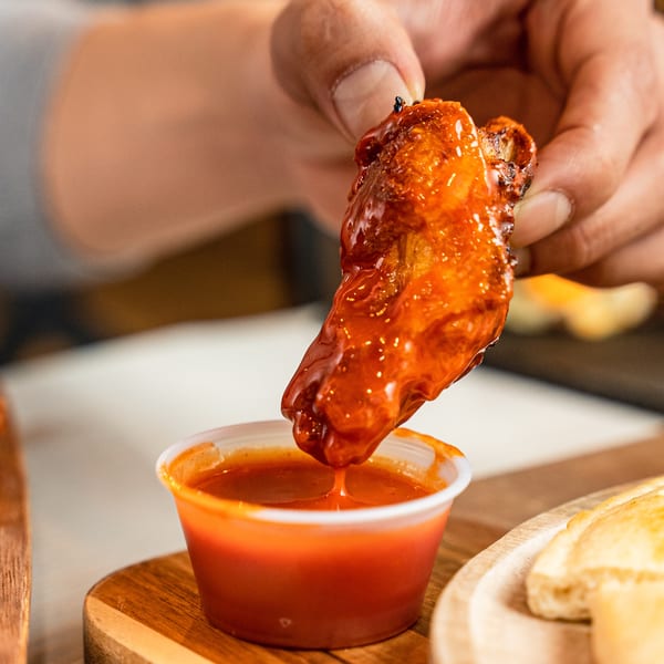 A hand dips a saucy, red chicken wing into a small cup of hot sauce.