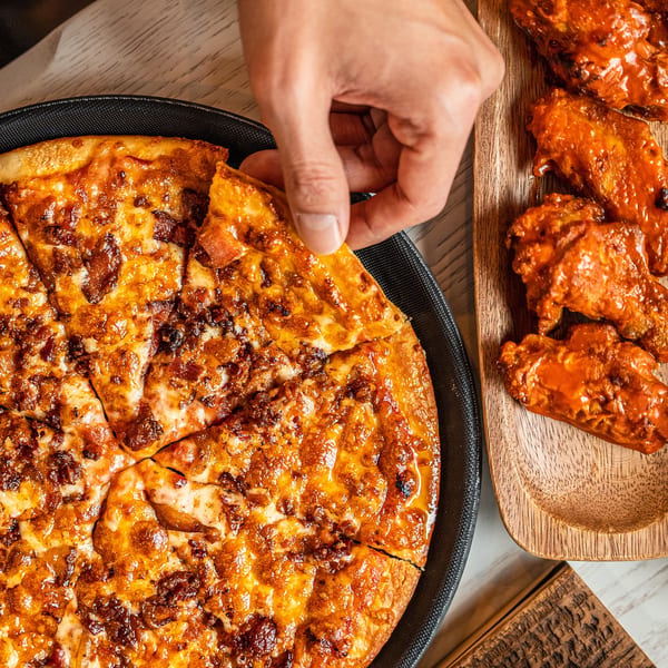 Hand grabbing a slice of Singas Famous Pizza with a side of buffalo wings.
