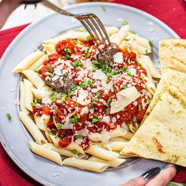 Penne pasta with meatballs, red sauce, parmesan, chives, and flatbread on a gray plate.