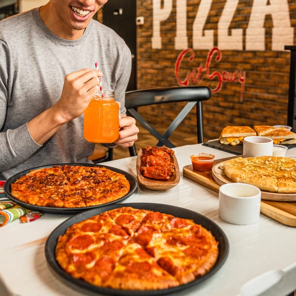 A person smiling, holding an orange drink, with two pizzas, wings, and garlic bread on a table.