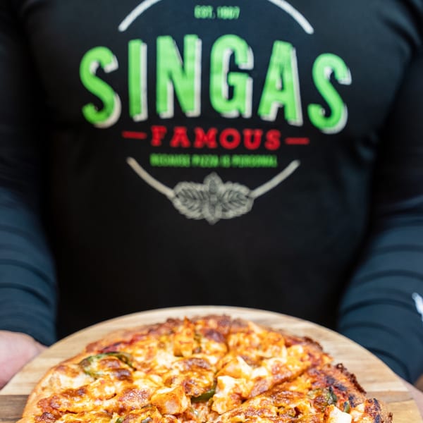 A person in a Singas Famous Pizza shirt holds a freshly baked pizza with chicken and jalapeños.