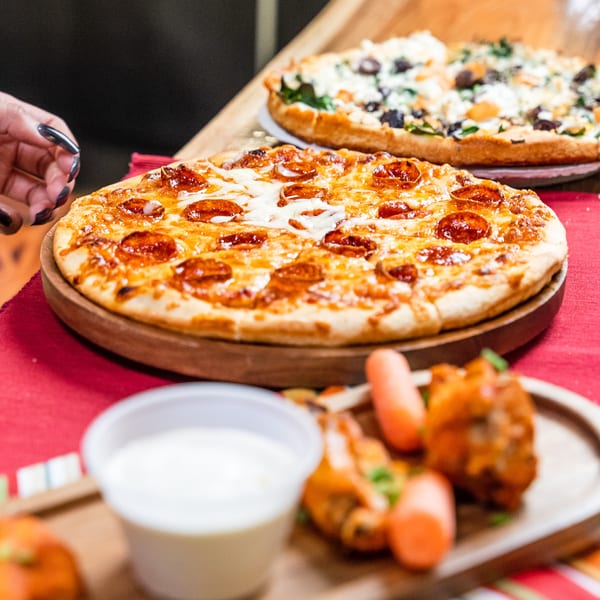A hand reaches for a pepperoni pizza, with another pizza and chicken wings in the background.