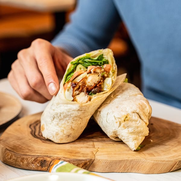 A hand holds a chicken and vegetable wrap, with another wrap resting on a wooden board.