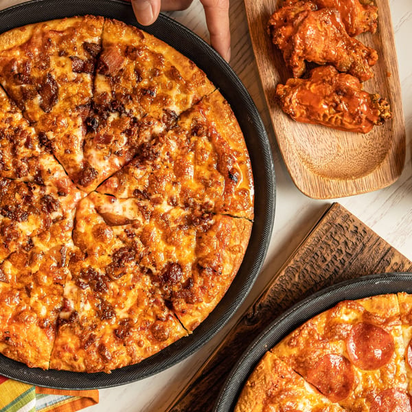 A hand serves a Singas Famous Pizza with a side of buffalo wings and another pepperoni pizza.