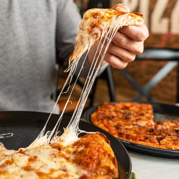 A hand pulls a slice of Singas Famous Pizza, creating long, gooey cheese strings.