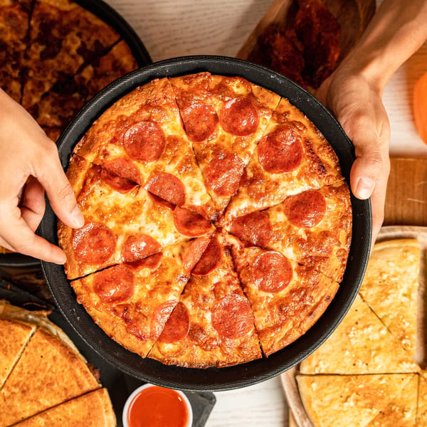 Hands holding a hot, sliced pepperoni pizza in a black pan, with other pizzas in the background.