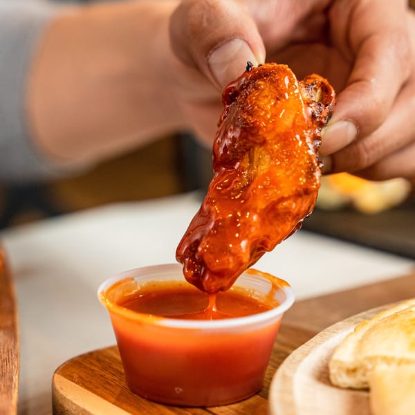 A hand dips a saucy chicken wing into a small plastic cup of red sauce.
