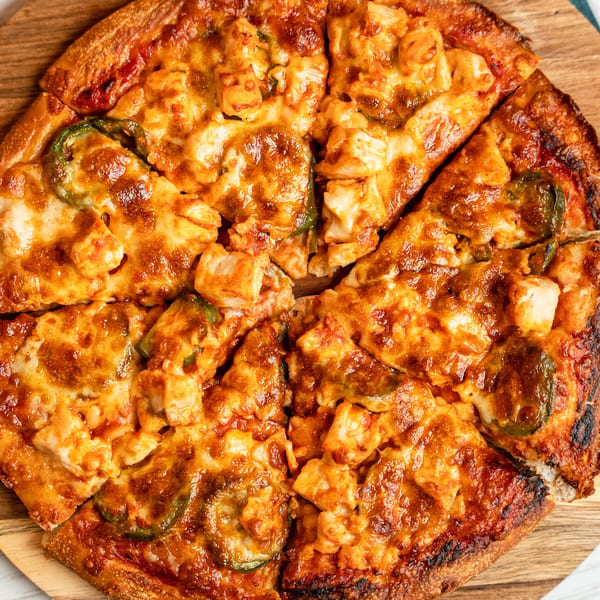 A close-up of a sliced Singas Famous Pizza with chicken, cheese, and jalapeños on a wooden board.