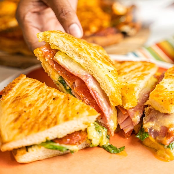 Hand picking up a slice of a golden-brown grilled sandwich filled with meat, cheese, and greens.