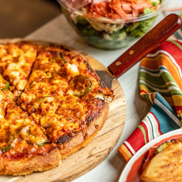 A sliced Singas Famous Pizza with melted cheese and toppings on a wooden board, next to a salad.