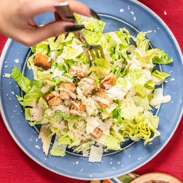 A hand with dark nails uses a fork to mix a Caesar salad with croutons and dressing on a blue plate.
