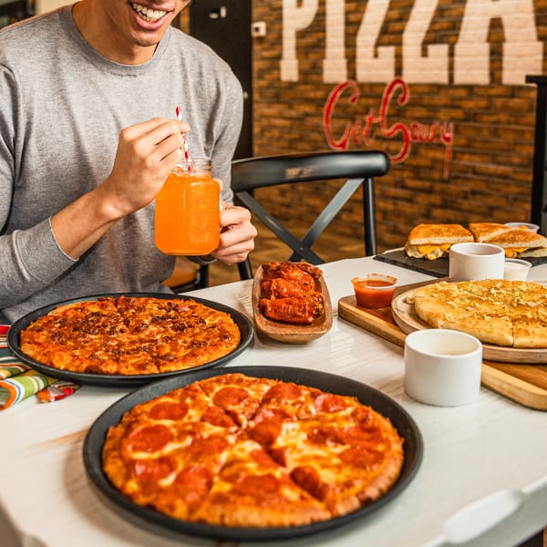 A person sips an orange drink at a table laden with Singas Famous Pizza, wings, and garlic knots.