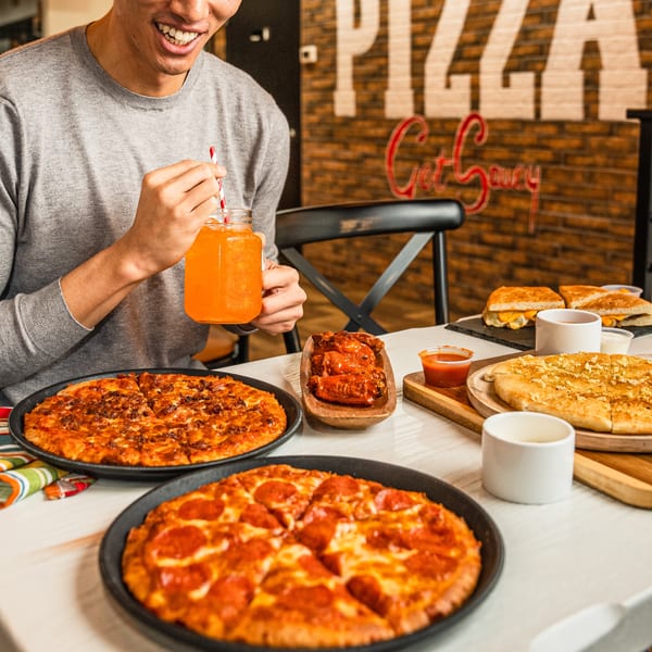 Smiling person enjoying orange juice with two pizzas, wings, garlic bread, and grilled cheese.
