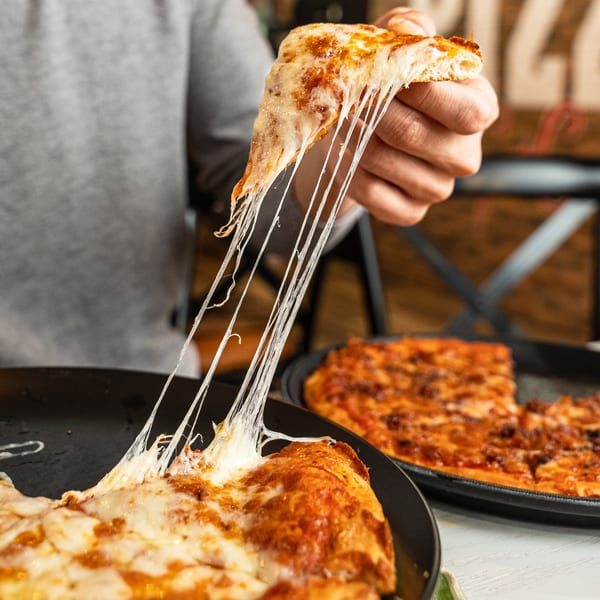 A hand pulls a slice of Singas Famous Pizza, stretching melted cheese, with another pizza in the background.