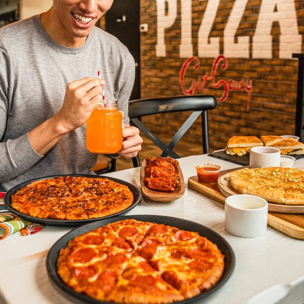 A person smiling, holding an orange drink, with two pizzas, wings, garlic knots, and grilled cheese on a table.