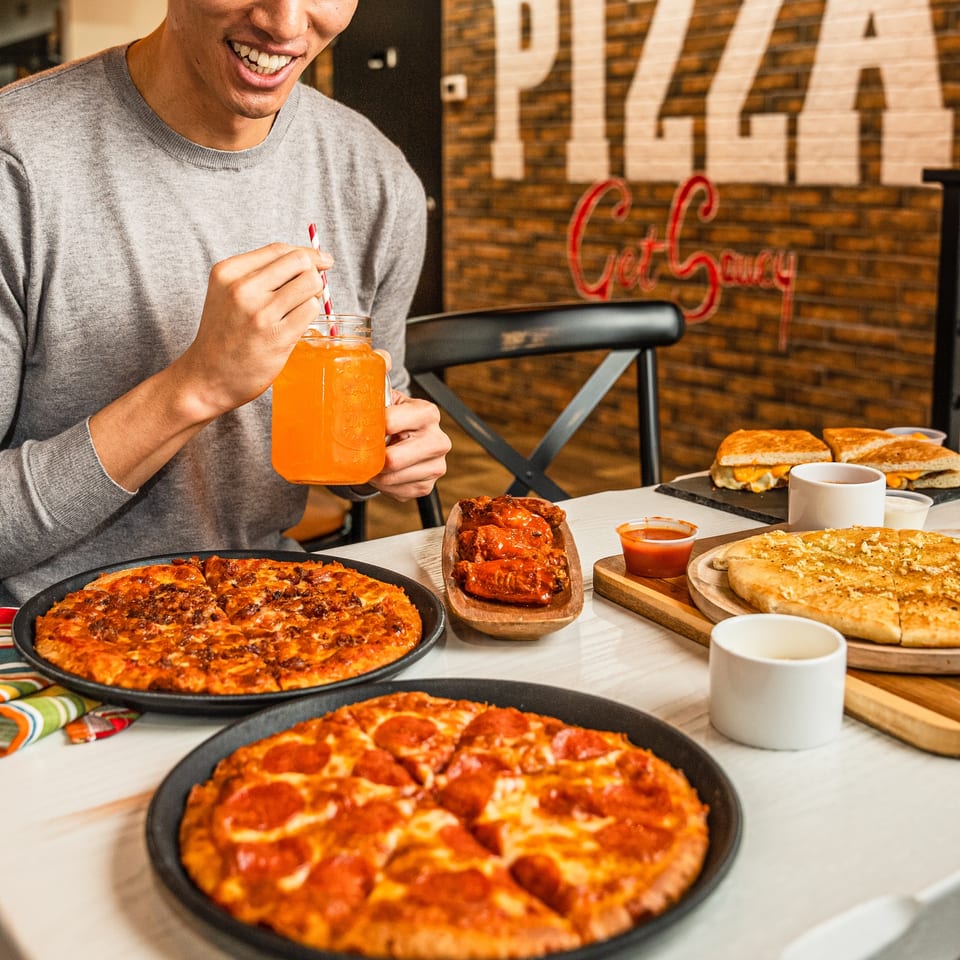 A person smiling, holding an orange drink, with two pizzas, wings, garlic knots, and grilled cheese on a table.