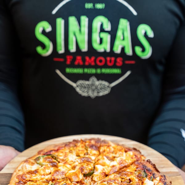 A person wearing a Singas Famous Pizza shirt holds a freshly baked pizza on a wooden peel.