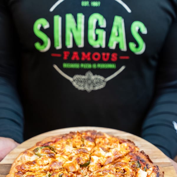 A person wearing a Singas Famous Pizza shirt holds a freshly baked pizza on a wooden peel.
