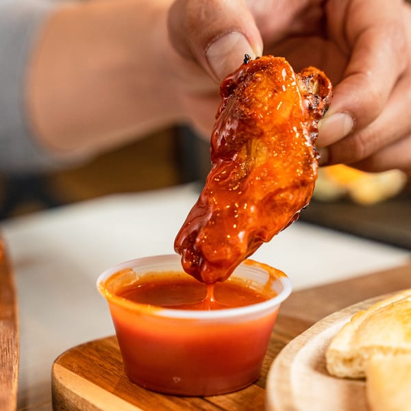 A hand dips a saucy chicken wing into a small cup of red dipping sauce.
