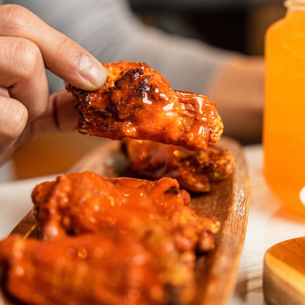 A hand picks up a saucy buffalo chicken wing from a wooden platter, with an orange drink nearby.