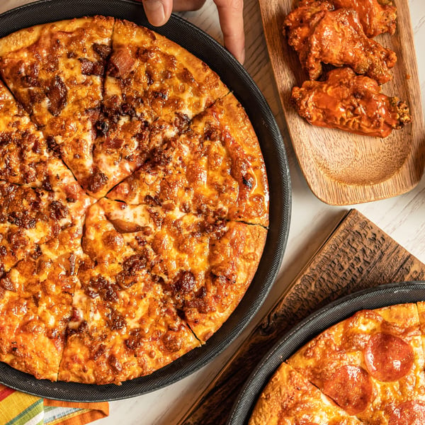 A hand serving a large sliced pizza with bacon, alongside buffalo wings and a pepperoni pizza.