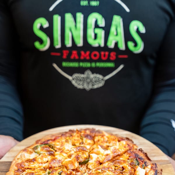 A person wearing a Singas Famous Pizza shirt holds a freshly baked pizza on a wooden peel.