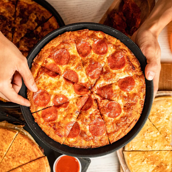 Hands holding a hot, sliced pepperoni pizza in a black pan, with other pizzas and sauce nearby.