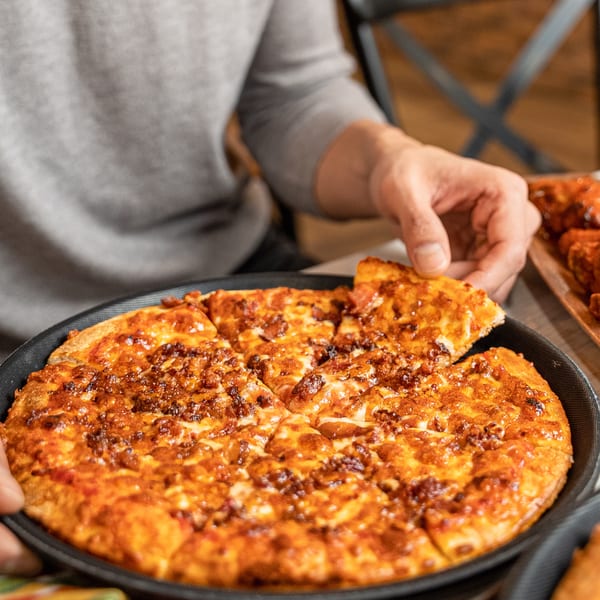 A person's hand reaching for a slice of a freshly baked, cheesy Singas Famous Pizza.