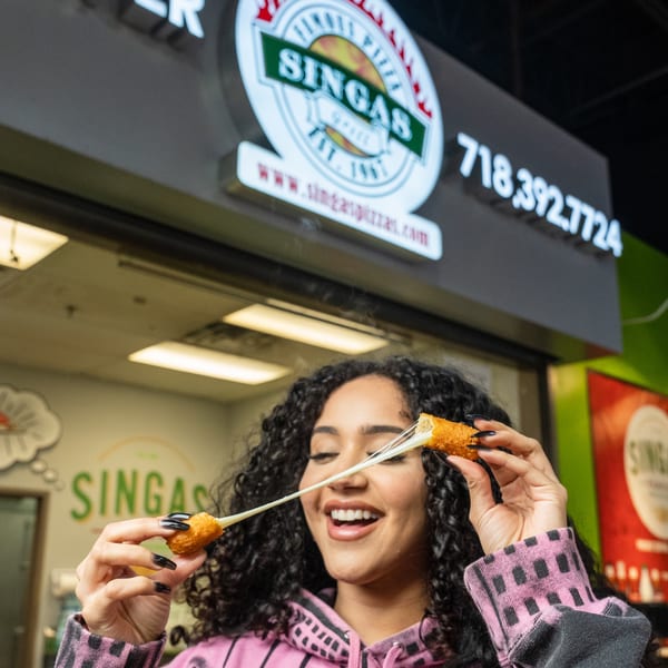 A smiling woman stretches cheesy mozzarella sticks in front of a Singas Famous Pizza sign.
