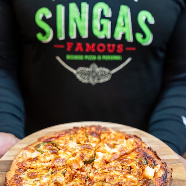 A person wearing a Singas Famous Pizza shirt holds a freshly baked chicken and pepper pizza.