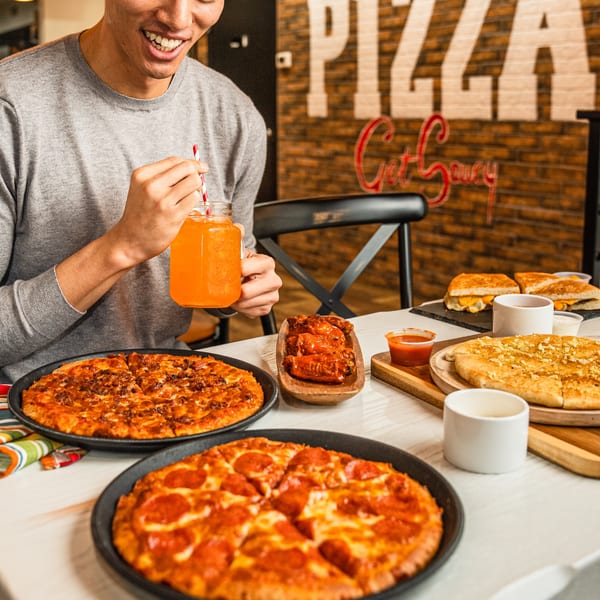 A person enjoying Singas Famous Pizza with two pizzas, wings, garlic bread, and a drink on a table.