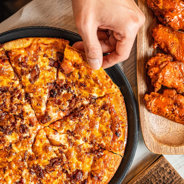 A hand grabbing a slice of Singas Famous Pizza with a side of buffalo wings.