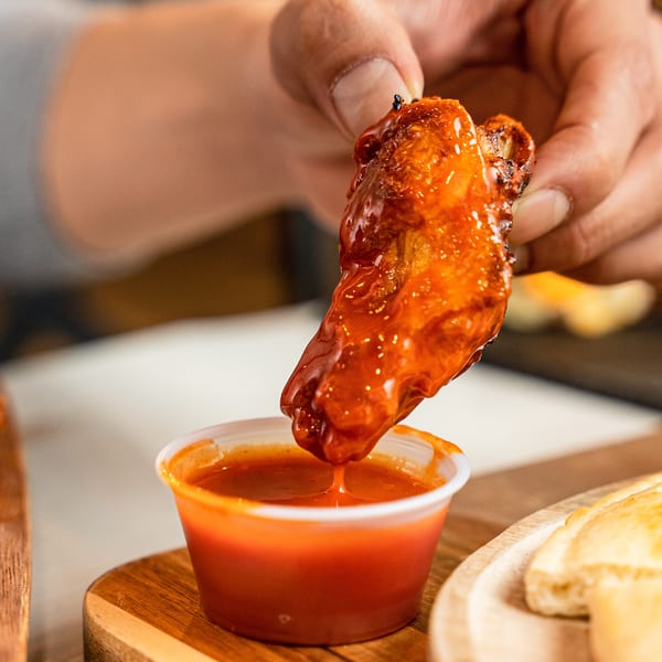 A hand dips a saucy chicken wing into a small cup of red dipping sauce.