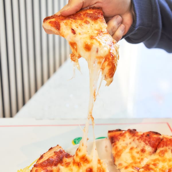 A hand holds a slice of Singas Famous Pizza with long, stretchy melted cheese.
