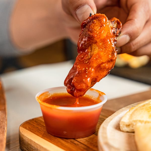 A hand dips a saucy chicken wing into a small cup of red dipping sauce.