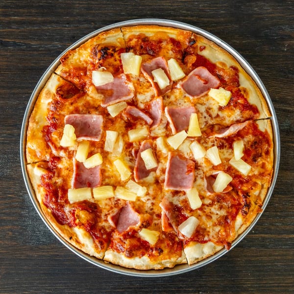 Singas Famous Pizza's Hawaiian pizza, topped with ham and pineapple, on a dark wooden table.