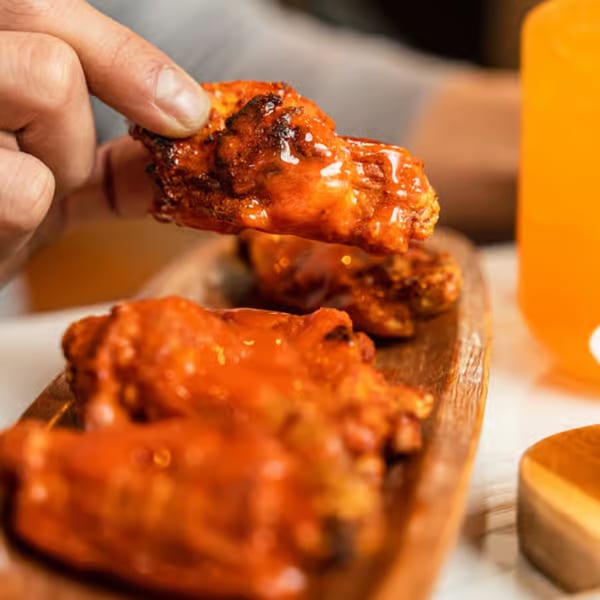 A hand picks up a saucy buffalo chicken wing from a wooden platter at Singas Famous Pizza.