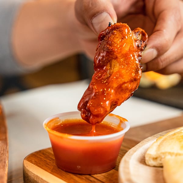 A hand dips a glossy, red-sauced chicken wing into a small cup of hot sauce.