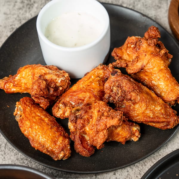 Crispy fried chicken wings on a black plate with a side of creamy white dipping sauce.