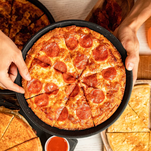 Hands holding a freshly baked, sliced pepperoni pizza in a black pan, with other pizzas in the background.