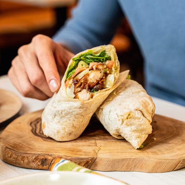 A person holding a chicken wrap filled with lettuce, tomatoes, and sauce on a wooden board.